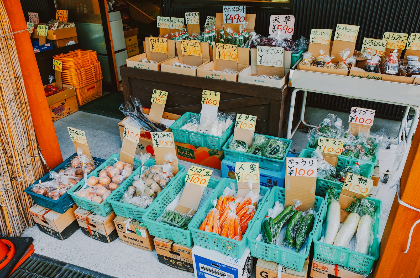 プラスチックのコンテナと段ボール箱に並んだ野菜に、手書きの値段札が立てられた直売所の店先